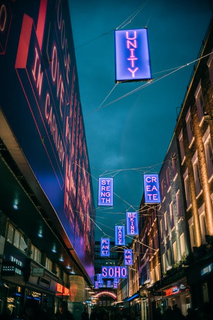 A vibrant nighttime scene on Carnaby Street in Soho W1, featuring bustling pedestrians walking along a cobblestone street illuminated by streetlights and colorful neon signs. The street is lined with 