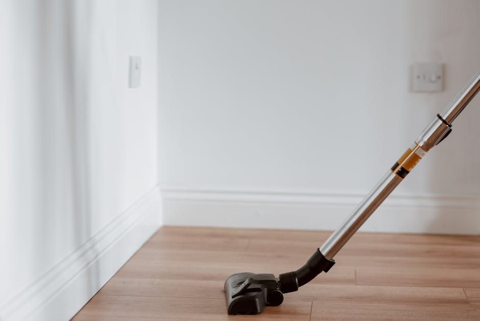 Close-up of a modern handheld vacuum cleaner with a metallic and black plastic body, resting on a light wooden floor adjacent to a white skirting board and plain white wall. The vacuum's transparent d